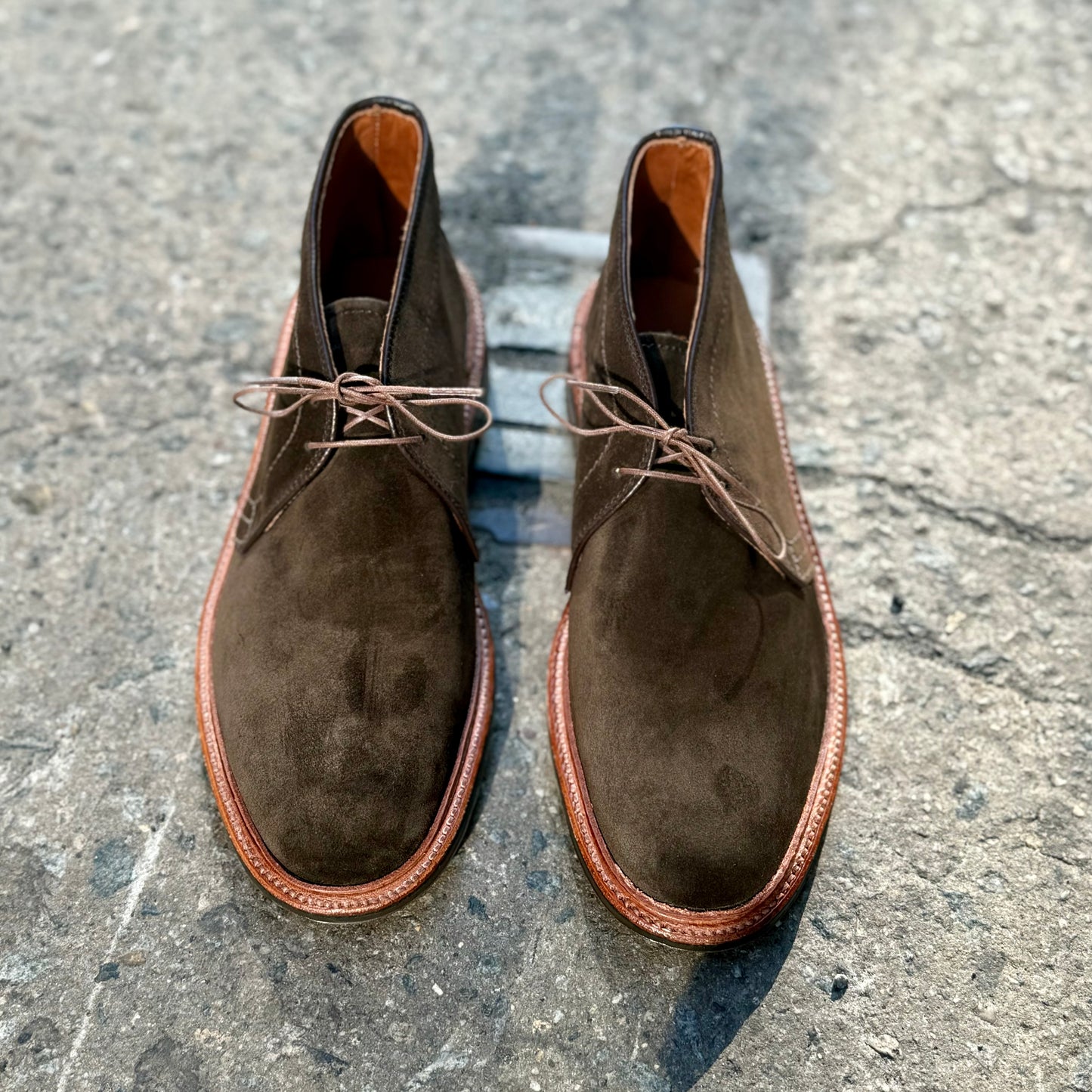 A pair of Alden Madisons D1712L Chukka Boots with L3 Sole in Loden Suede sits on a textured gray concrete surface. Featuring the Barrie Last design, these boots have round toes and neatly tied brown laces. Lighting casts a subtle shadow around them, highlighting their craftsmanship.