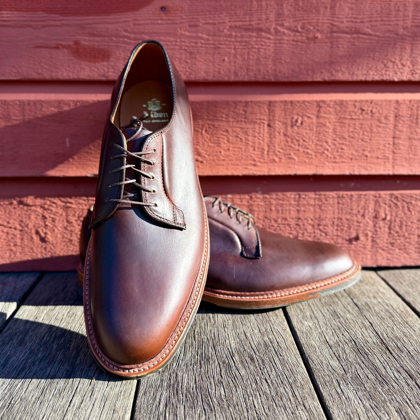 A pair of ALDEN SHOES 9506C Plain Toe Blucher w Commando Brown CXL is shown outdoors on wooden planks before a red wall; one Barrie Last shoe stands upright while the other lies on its side behind it.