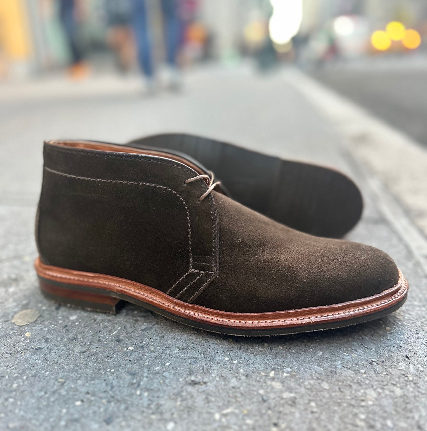 The Alden Madison D1712L Chukka Boot in Loden Suede elegantly rests on a city sidewalk, showcasing its craftsmanship with an L3 Sole while the vibrant urban backdrop adds a dynamic blur.