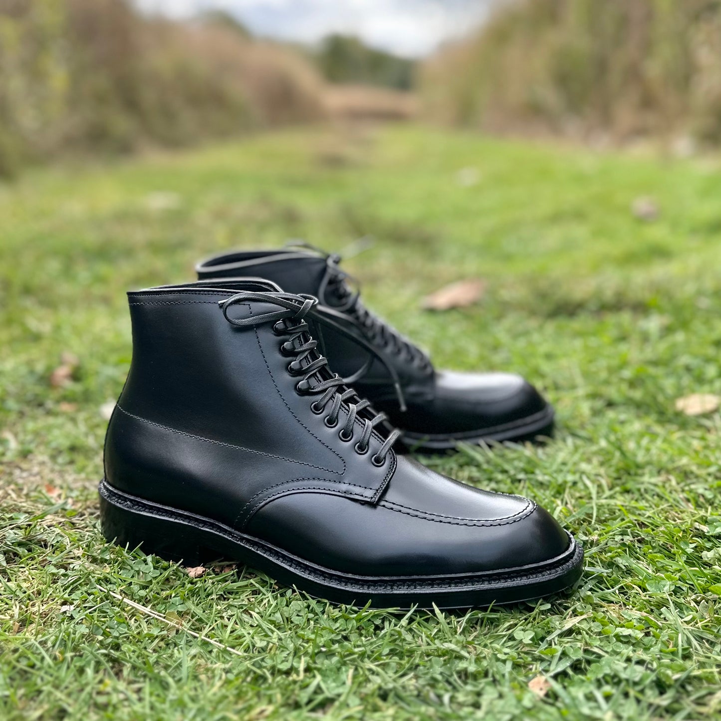 A pair of Alden Madisons D2941H - Black Calf Indy Boots, with a double leather sole and laces, rests on a grassy field. The blurred greenery in the backdrop highlights their exquisite craftsmanship and sleek design.