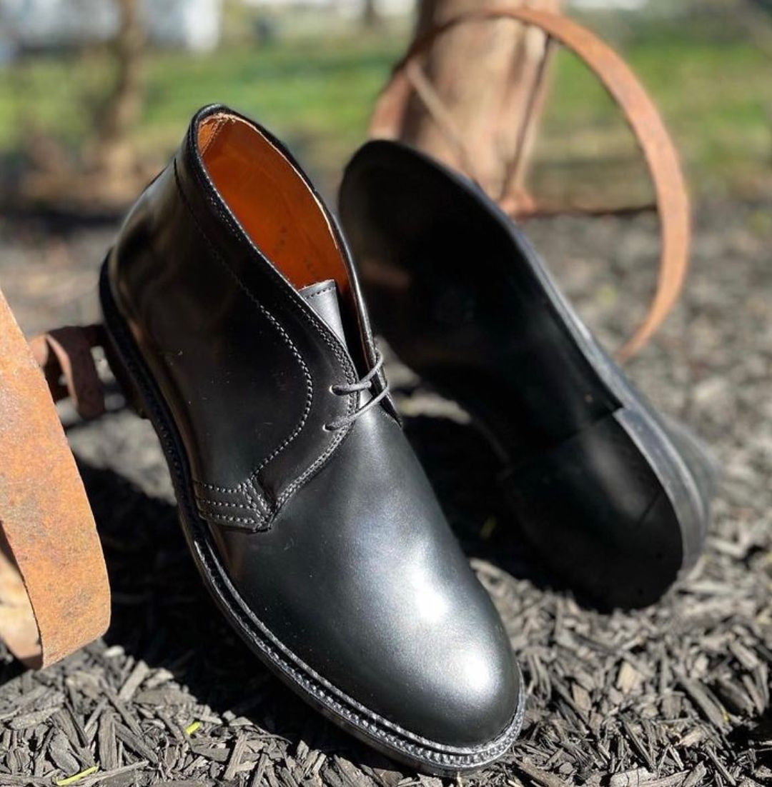 A pair of Alden Madisons 1340 - Chukka Boot in Black Shell Cordovan sits outdoors on dark mulch, with a rusty iron wheel and blurred greenery in the background, enhancing the rustic charm.