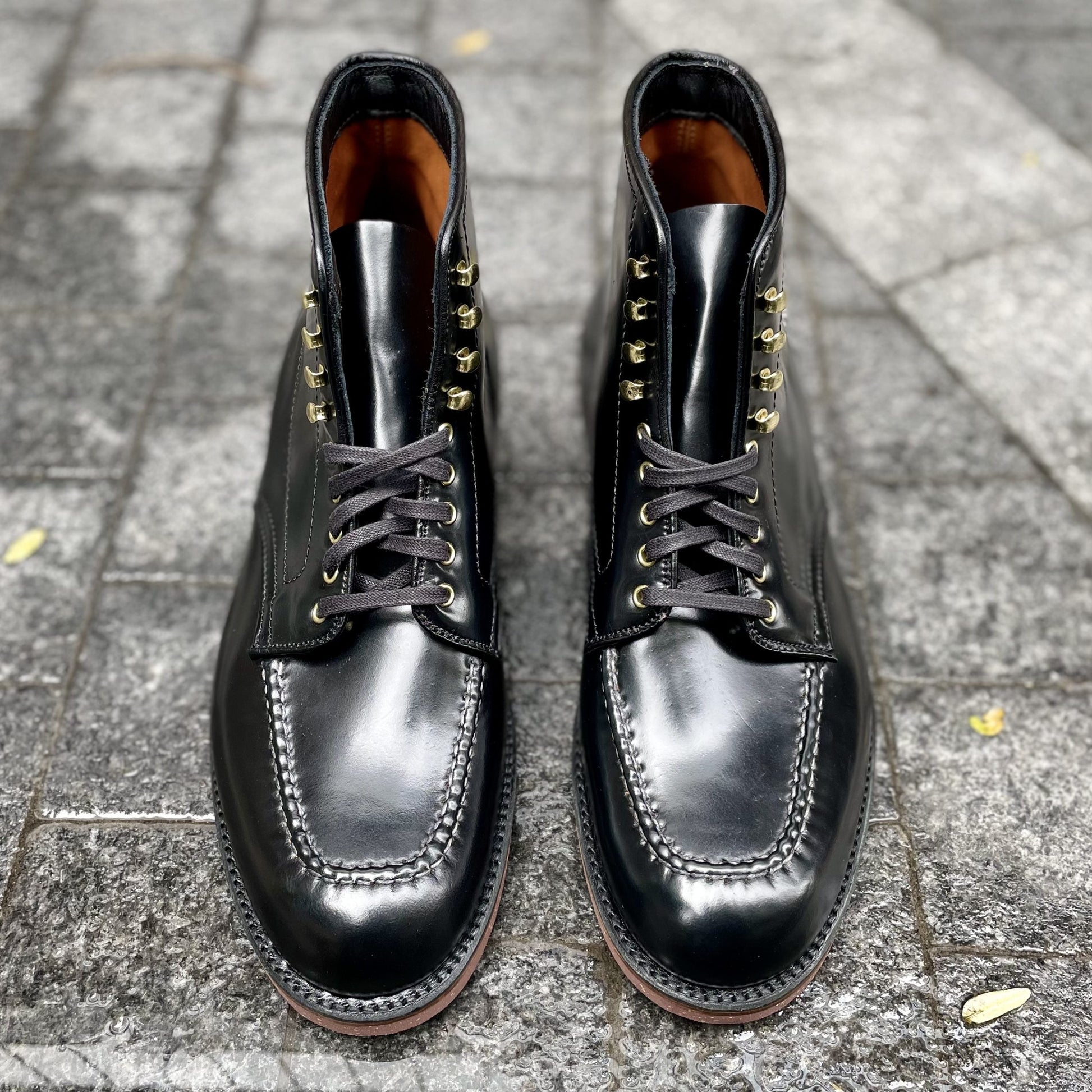 The Alden Madison D1908H Copake Indy Boot in Black Shell Cordovan features detailed hand stitching and gold eyelets, displayed side by side on a textured stone pavement.