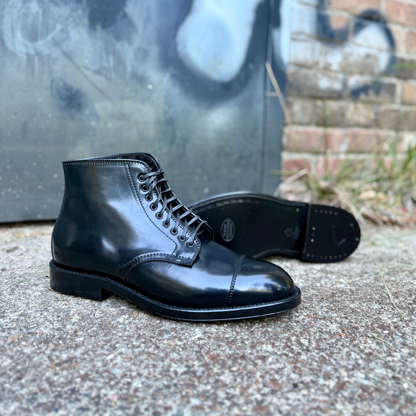 The Alden Madison 4065 Cap Toe Boot in Black Shell Cordovan, featuring a Double Oak Leather Sole, is displayed outdoors on rough concrete with one boot upright and one on its side; a brick wall and graffiti-covered metal door are in the background.