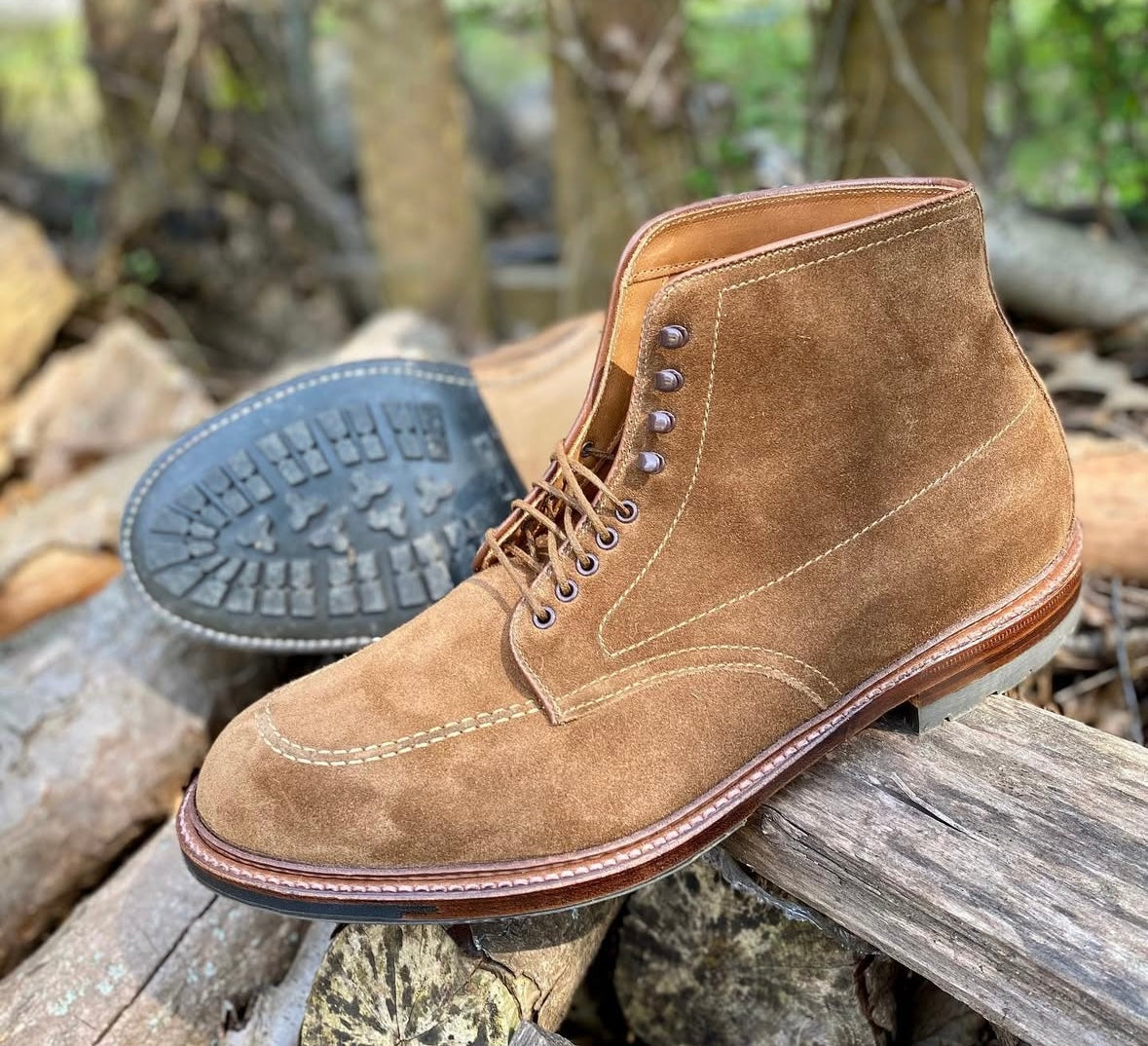 The Alden Madison 4011HC - Indy Boot in Snuff Suede features visible stitching and an antique commando sole, shown outdoors on stacked logs with greenery softly blurred in the background.