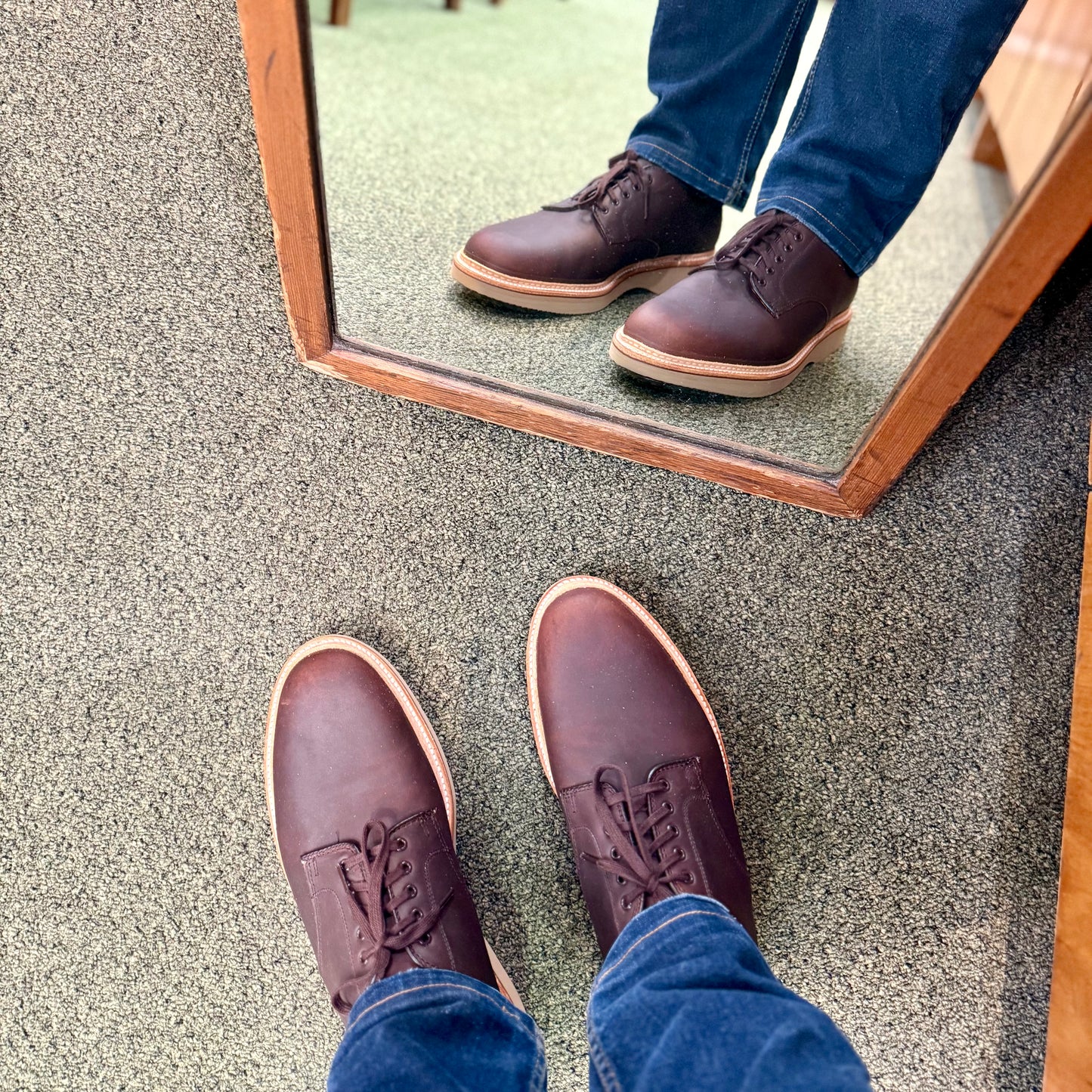 A person wearing Alden Madison 29401 - The “Ruslan” Plain Toe Dover in Kudu leather with a tan wedge rubber sole and blue jeans stands on a carpeted floor before a mirror, with their shoes and jeans reflected.