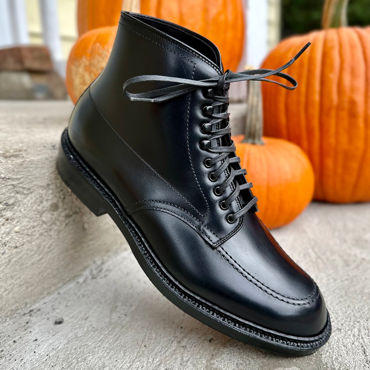 The D2941H - Black Calf Indy Boot by Alden Madison, featuring detailed stitching and laces, rests on a concrete step with three orange pumpkins in the background for an autumnal touch.