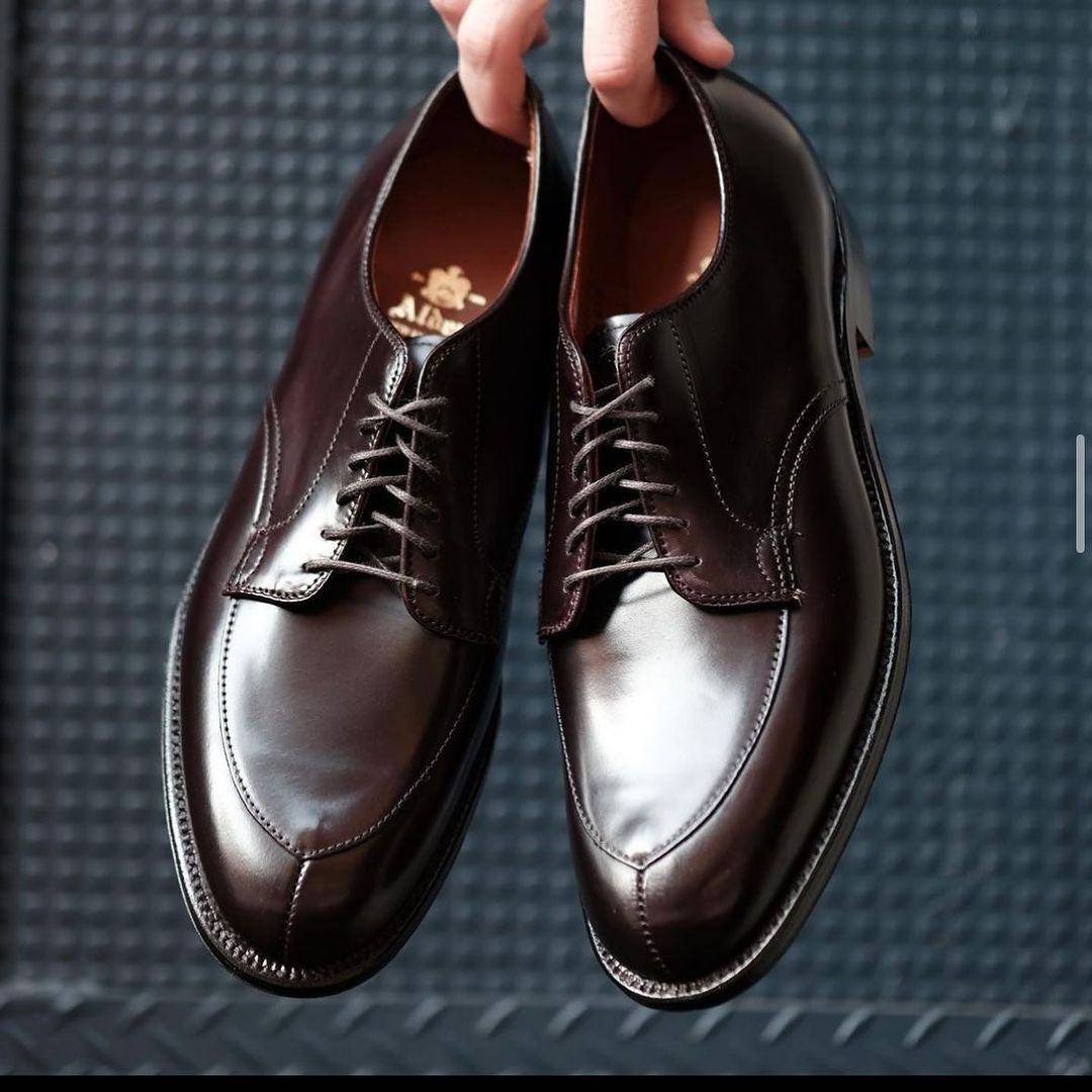 The Alden Madison D6616 V-Tip Split Toe Blucher in Color 8 Shell Cordovan, with a polished finish and visible stitching, dangle by their laces against a textured dark background, revealing slightly visible soles.