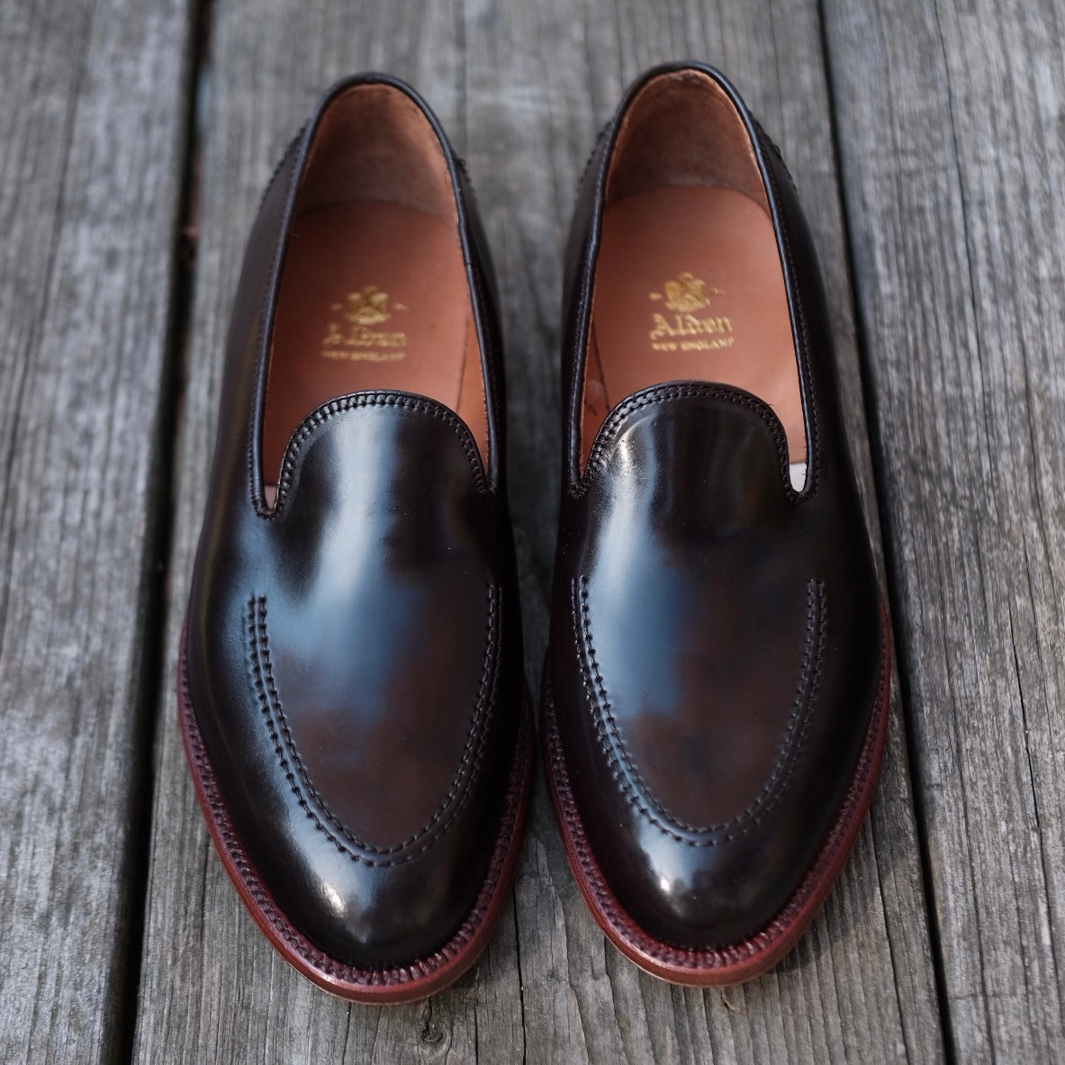 A pair of Alden Madisons D1118 - Vanderbilt Slip Ons in deep brown shell rests on a wooden surface, adorned with detailed stitching. The gold-text embossed insoles and the polished finish on the Aberdeen Last highlight their impeccable craftsmanship and reflect light brilliantly.