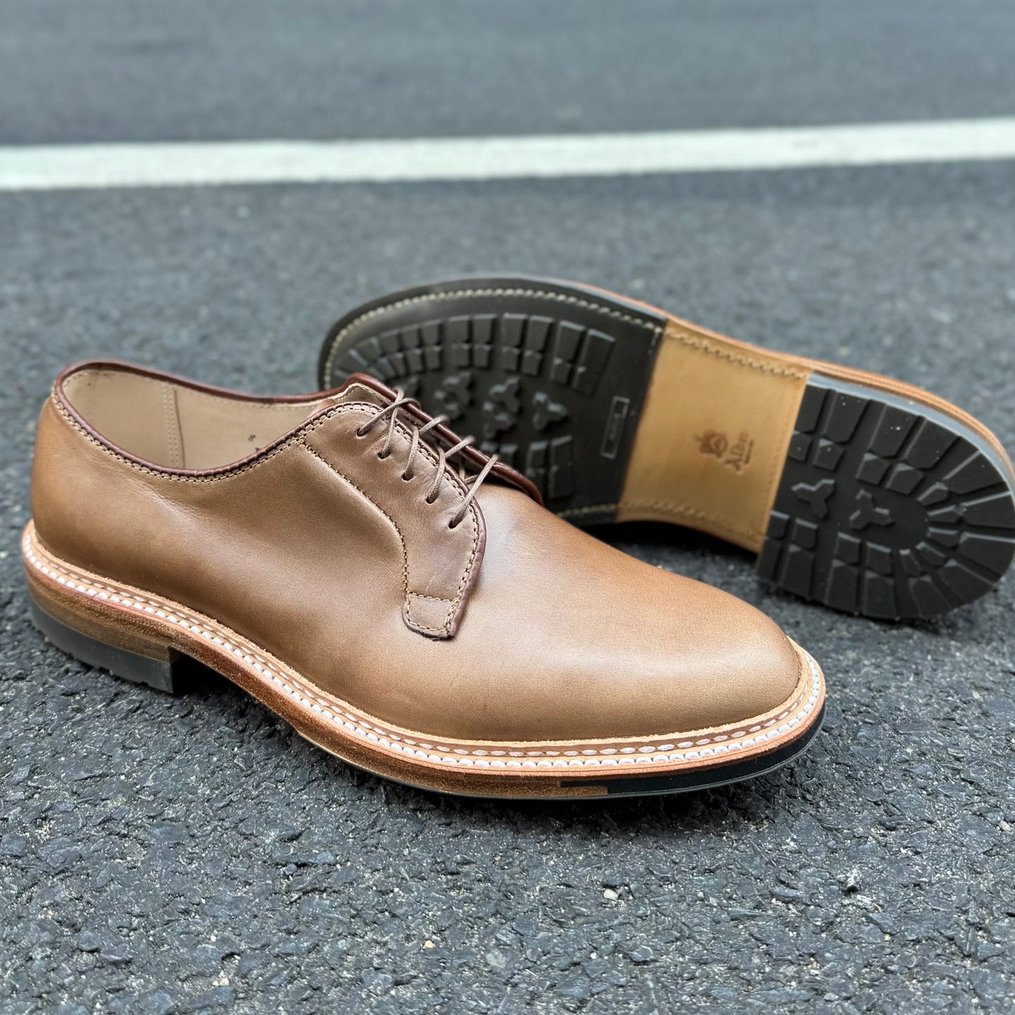 A pair of ALDEN SHOES 9501C - Natural CXL Plain Toe Blucher dress shoes with visible stitching sit on asphalt; one stands upright while the other displays its black rubber sole.