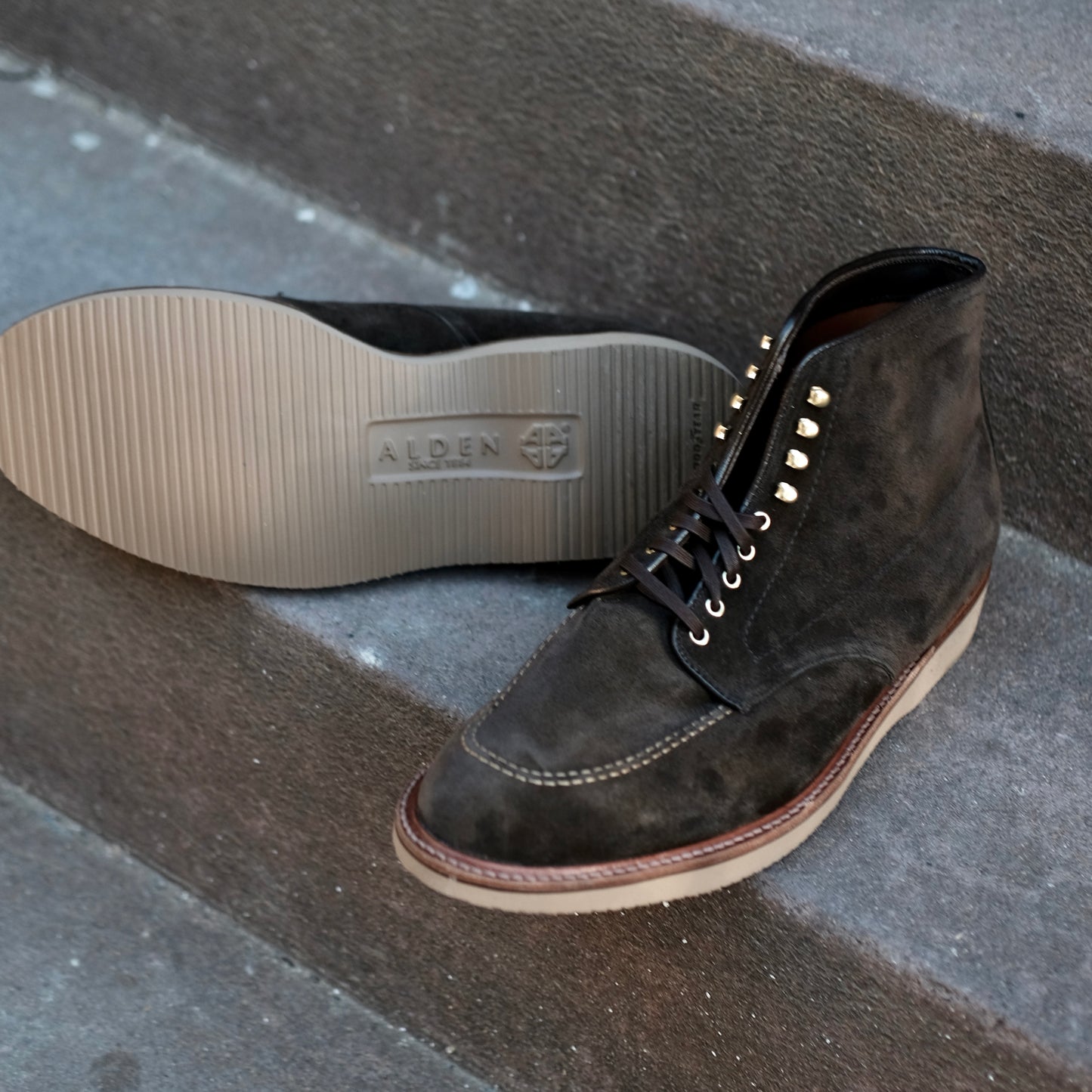 A pair of Alden Madison D0938H Duncan Indy Boots in Loden Suede with light rubber soles, one upright and one on its side showing the ALDEN logo, displayed on concrete steps.