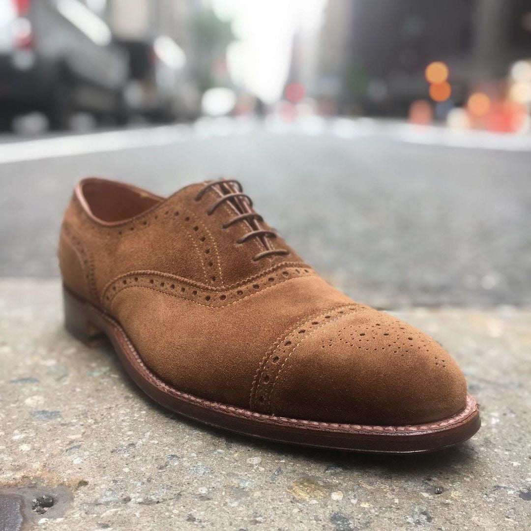 The Alden Madison 51670F Medallion Cap Toe Bal in Snuff Suede, crafted on the Hampton Last with brogue details and a Flex leather sole, sits on a city street with a blurred urban background and faintly visible cars.