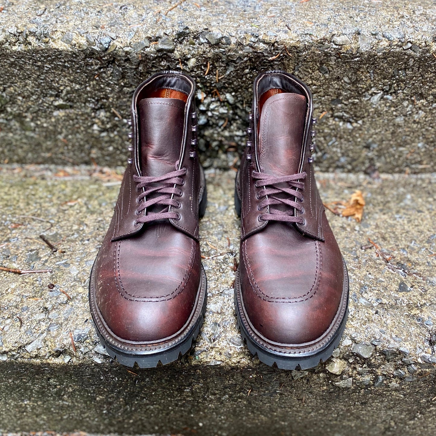 A pair of Alden Madisons 404 - Indy Boot in Kudu (Oiled CXL) rests on concrete steps. Featuring rugged kudu leather, Vibram lug outsoles, and metal eyelets, they blend durability with style. The steps beneath showcase a rough, weathered look.