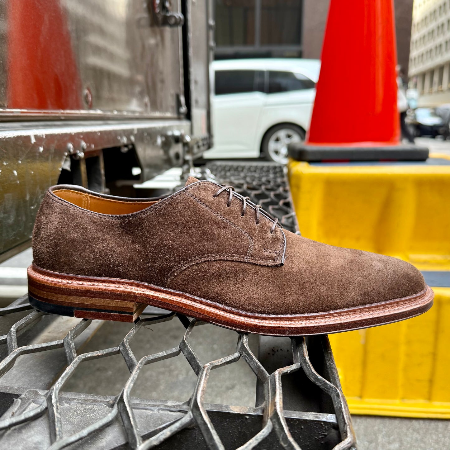 The Alden Madison 29335F - Unlined Dover in Brown Suede, crafted on the Barrie Last with a Flex Leather Sole, is displayed on a metal grate in an urban scene featuring a traffic cone and yellow barrier in the background.