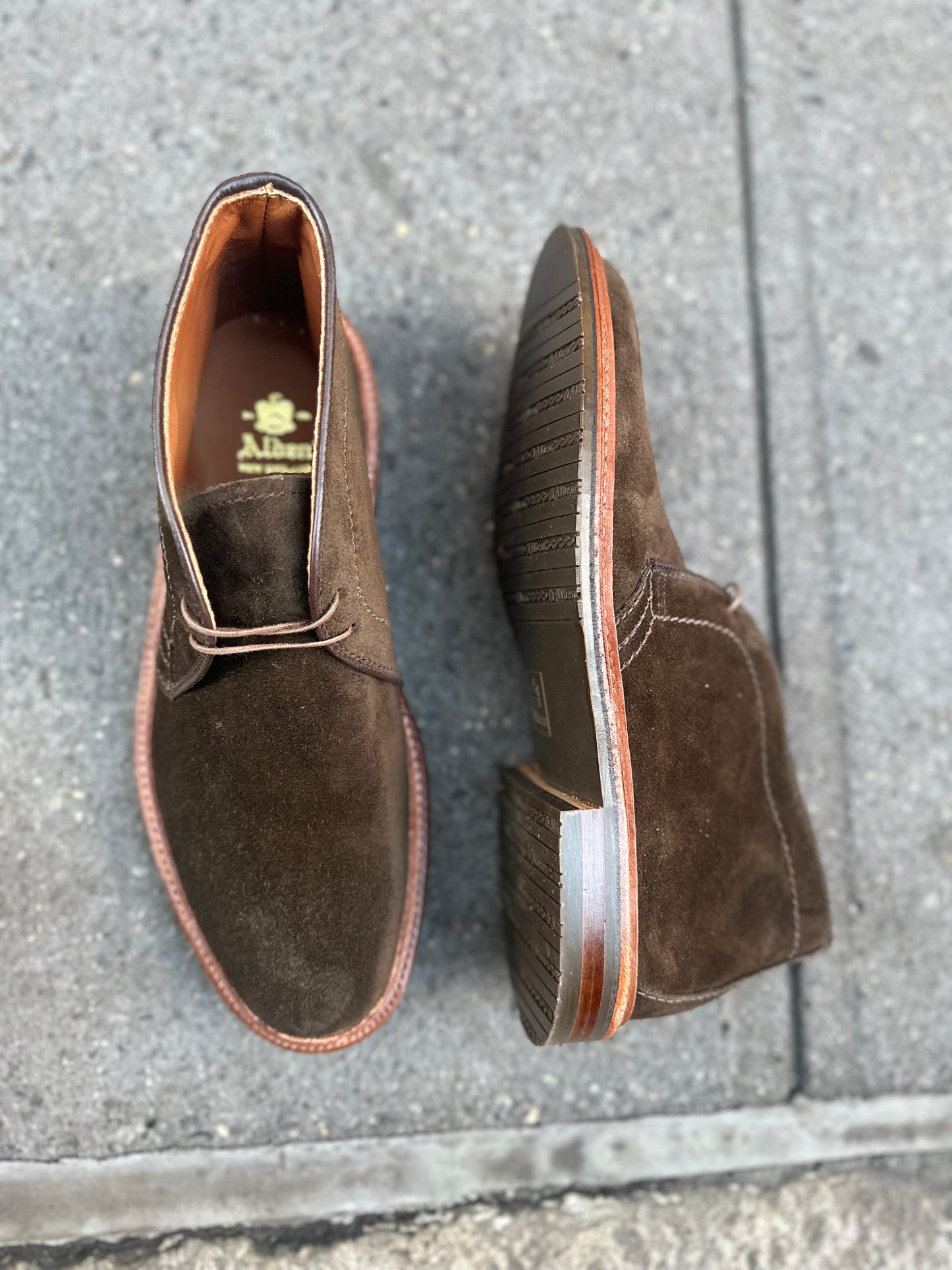 The Alden Madison D1712L Chukka Boot in Loden Suede, featuring an L3 Sole and crafted on the Barrie Last with a reverse welt, is showcased on gray textured pavement. One boot stands upright while the other lies on its side, revealing its intricate sole design.