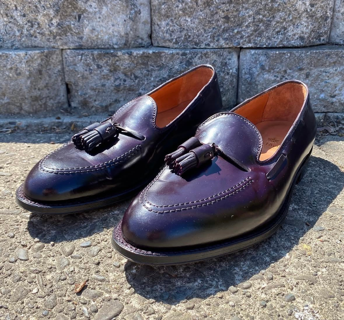 The Alden Madison 563 - Tassel Loafer in Color 8 Shell Cordovan, featuring a polished finish and sleek leather outsole, sits on concrete before a stone wall with sunlight gleaming off its rich dark brown exterior.
