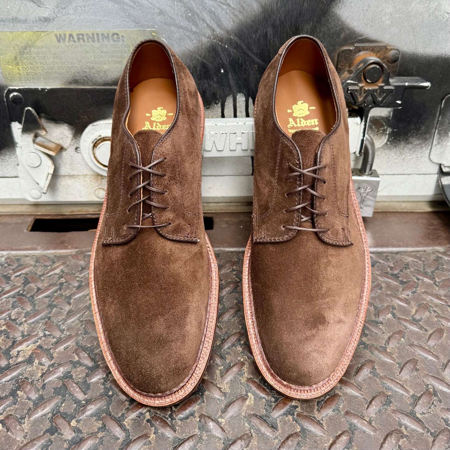 A pair of Alden Madison 29335F - Unlined Dover in Brown Suede with Flex Leather Sole rests on a textured metal surface before industrial equipment, featuring gold A. Albert branding on the insoles.