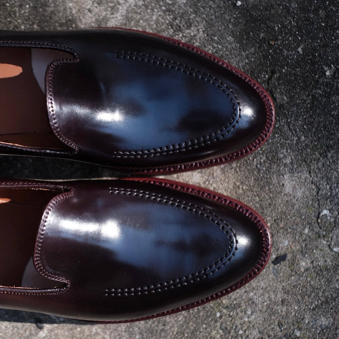 The Alden Madison D1118 - Vanderbilt Slip On in Color 8 Shell, a pair of shiny dark brown leather shoes with decorative stitching and maroon leather soles, sits on a gray concrete surface, reflecting light on their polished finish.
