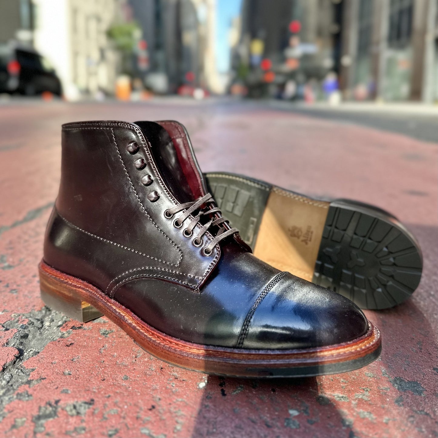 The Alden Madison D2824HC - Cap Toe Boot in Color 8 Shell, featuring a sturdy Commando sole, is displayed on an urban street. One boot stands while the other lies on its side, exposing the robust sole against a backdrop of blurred city buildings and cars.