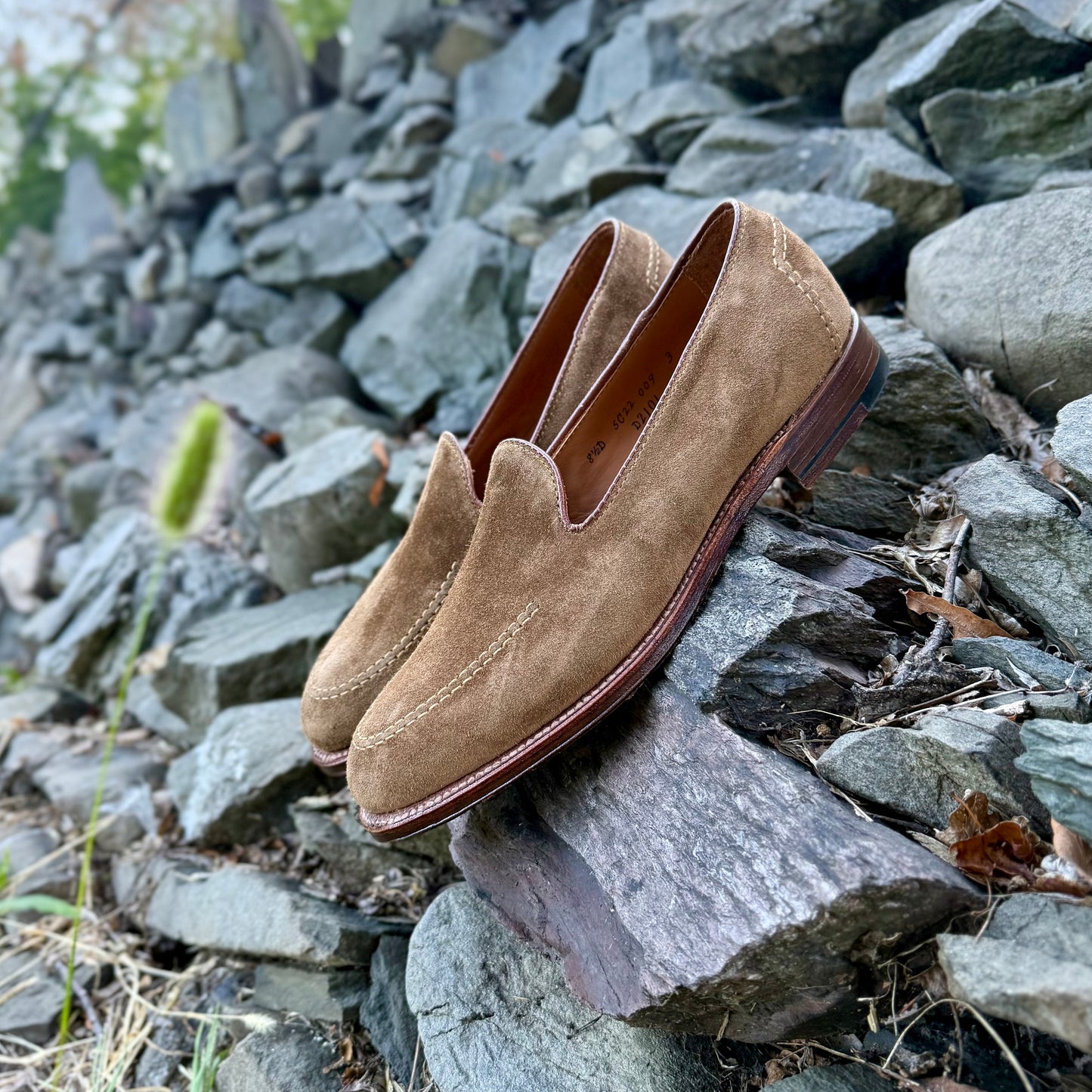 The D2101 - Vanderbilt Slip On in Snuff Suede by Alden Madison is displayed outdoors on gray, jagged rocks with greenery in the background.