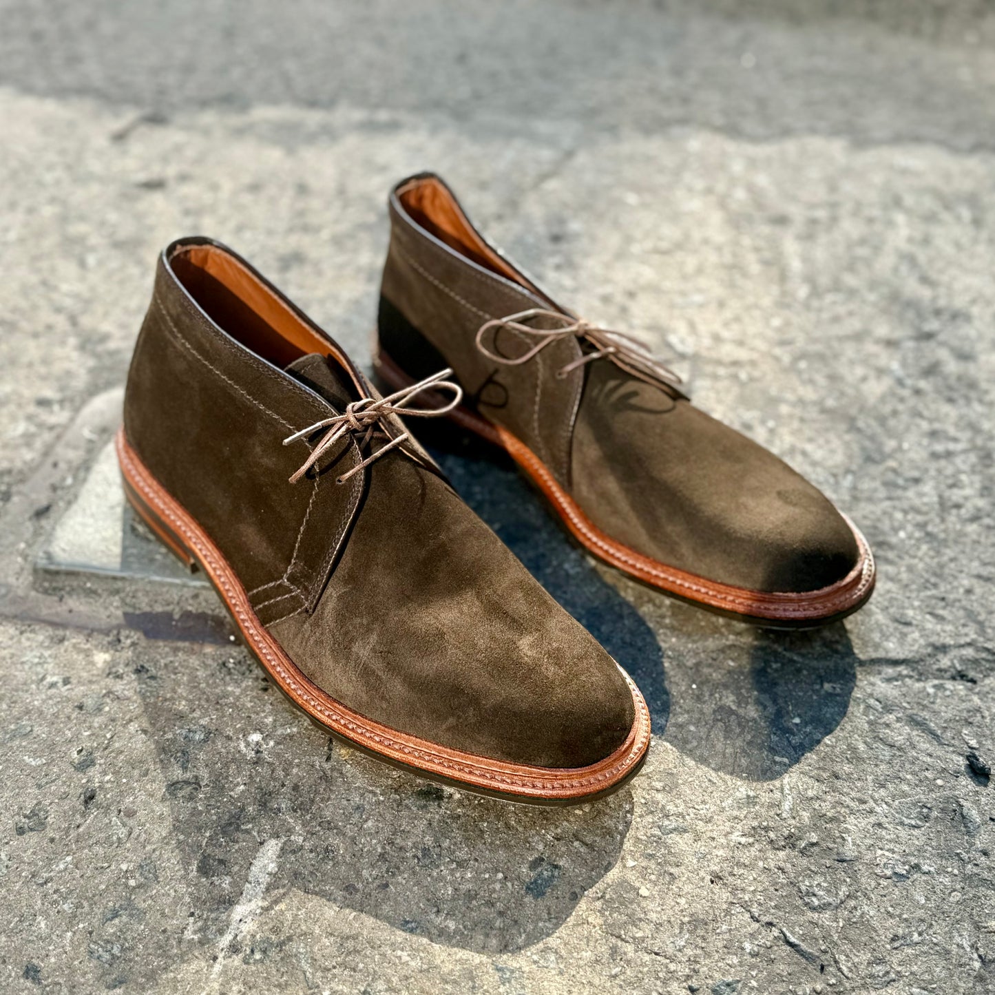 A pair of D1712L - Chukka Boot with L3 Sole in Loden Suede by Alden Madison, featuring a distinctive reverse welt and light brown laces, sits on a textured stone surface. The Barrie Last design offers exceptional comfort as soft sunlight casts gentle shadows around them.