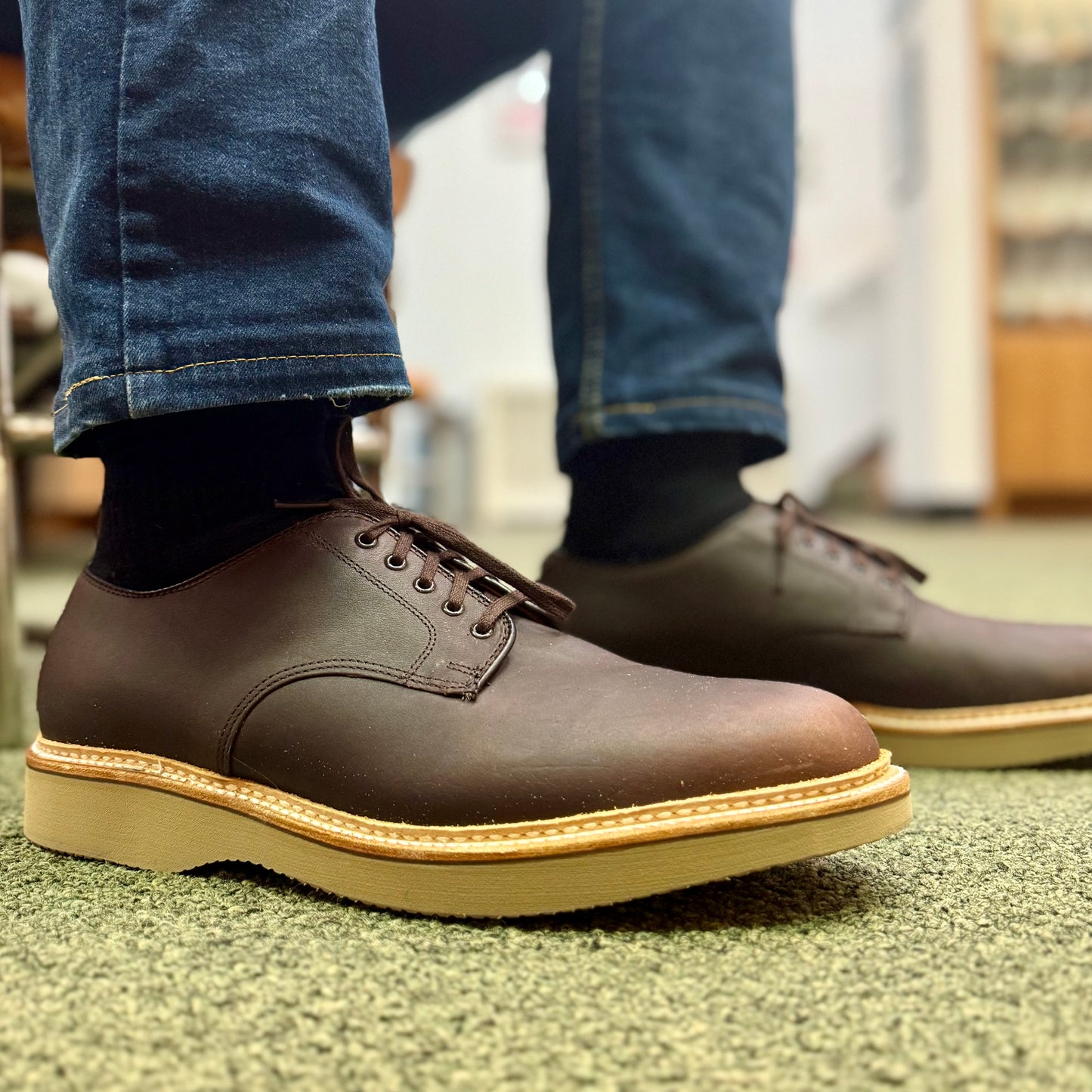 A close-up of a person wearing Alden Madison 29401 - The Ruslan Plain Toe Dover in Kudu Leather, with a tan wedge rubber sole, black socks, and blue jeans, standing on a green carpet indoors.