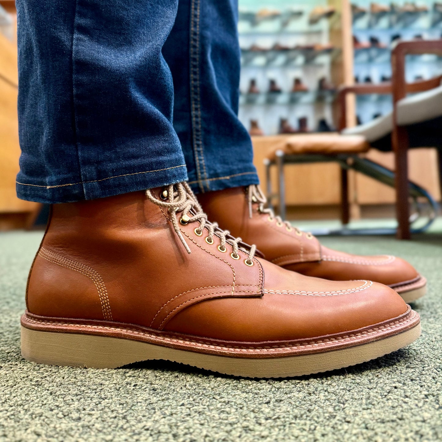 Someone stands on a carpeted floor wearing Alden Madisons Sherwood Indy Boot, made from Saddle Calf Leather with Trubalance Last and thick cream wedge soles. Theyre in blue jeans, and the store display behind them is blurred.