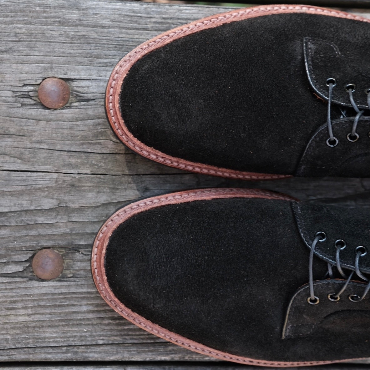 The D1426C - Plain Toe Blucher in Earth Reverse Chamois by Alden Madison features black suede with brown Commando soles and black laces, elegantly displayed on a weathered wooden surface with visible nails.