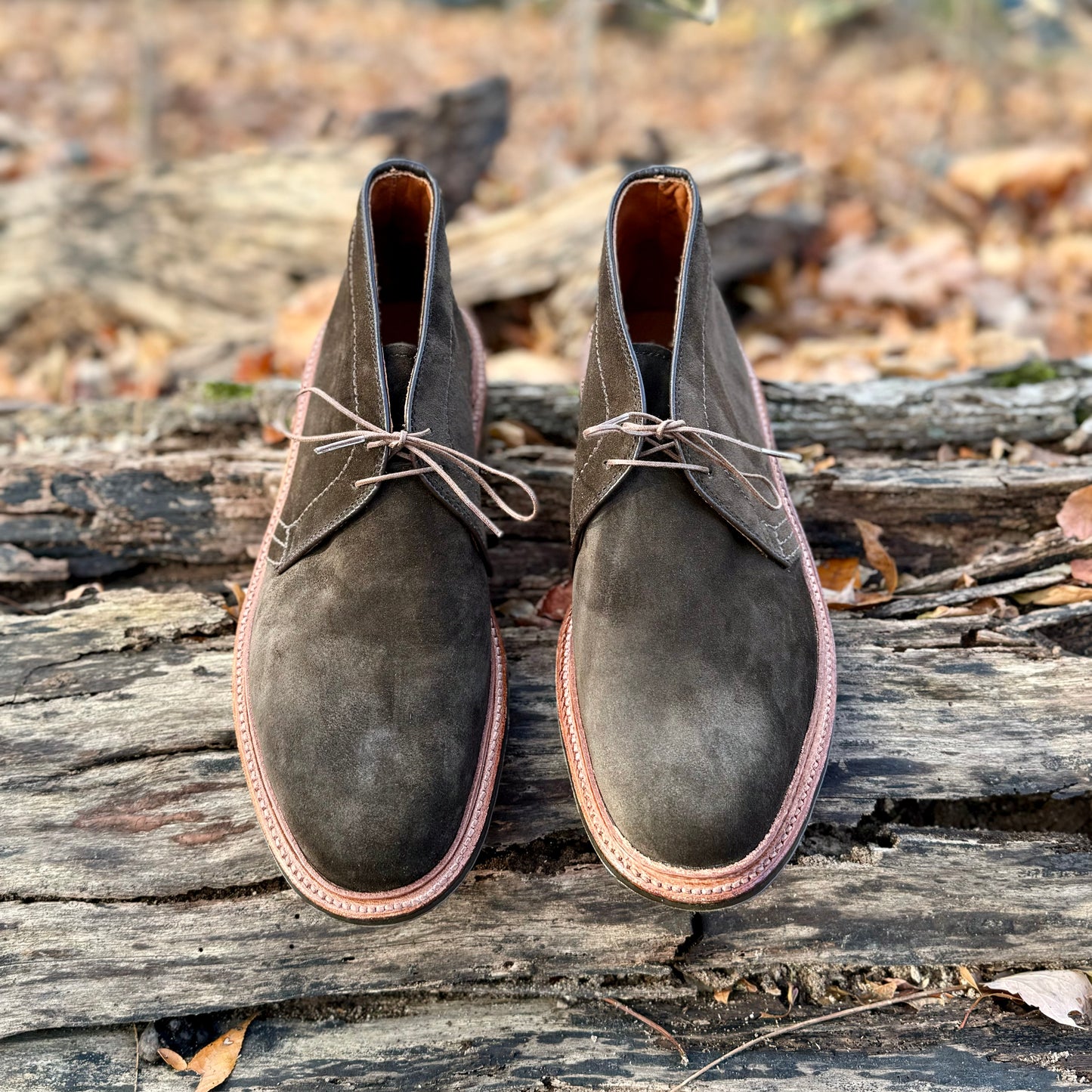A pair of Alden Madison D1712L Chukka Boots in Loden Suede, featuring L3 leather soles, laced up on a weathered wooden surface with autumn leaves evokes a rustic outdoor vibe.