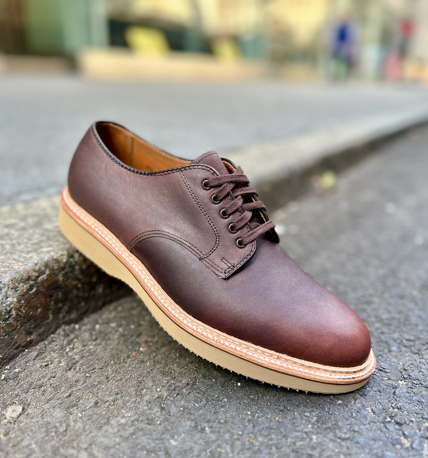 The Alden Madison 29401 Plain Toe Blucher in brown Kudu leather, featuring a light tan rubber sole and refined Barrie last shape, is angled on an outdoor concrete surface to highlight its clean stitching and sleek design.