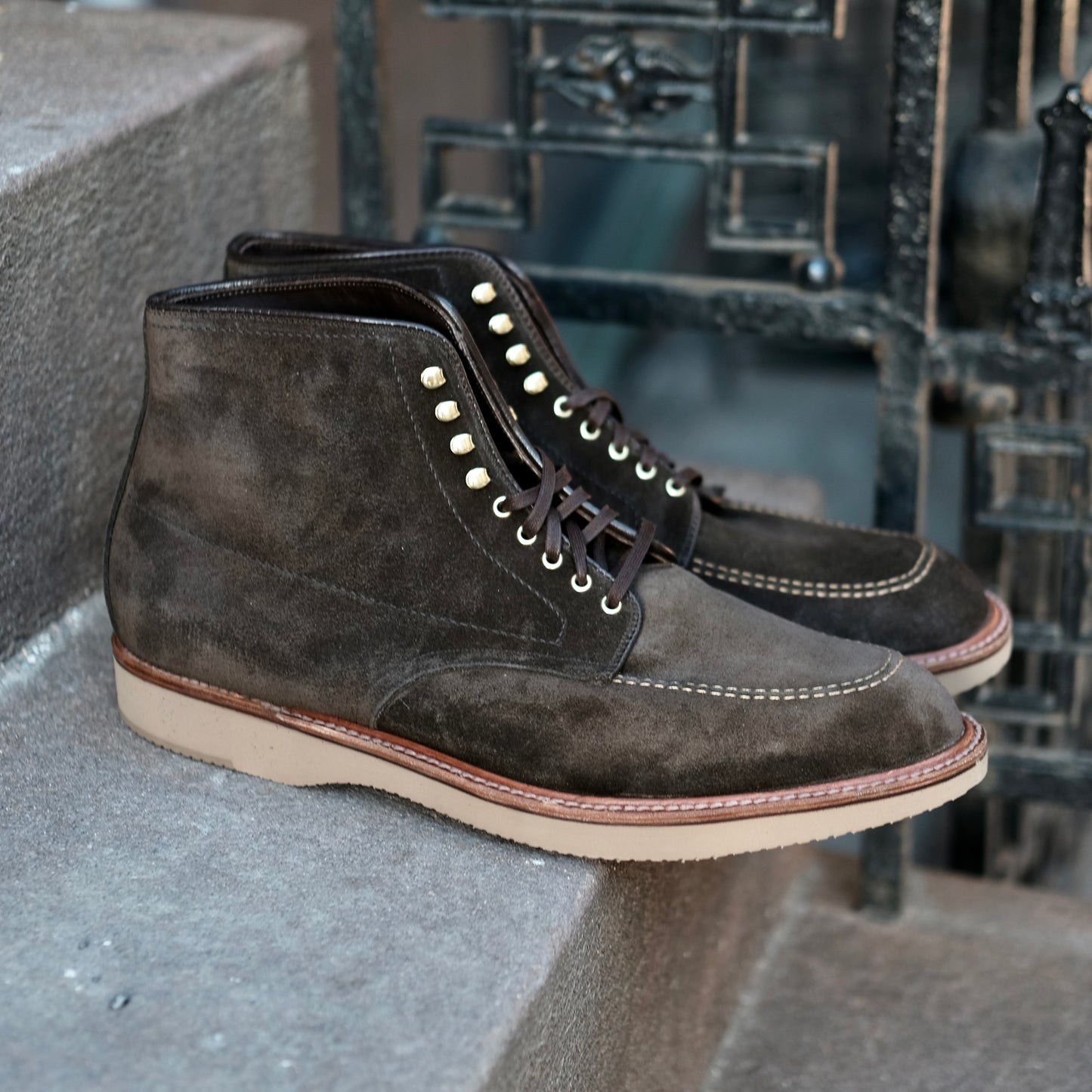 The Alden Madison D0938H - Duncan Indy Boot in Loden Suede, featuring light soles and built on the Truebalance Last, is shown on stone steps beside an ornate black metal railing.
