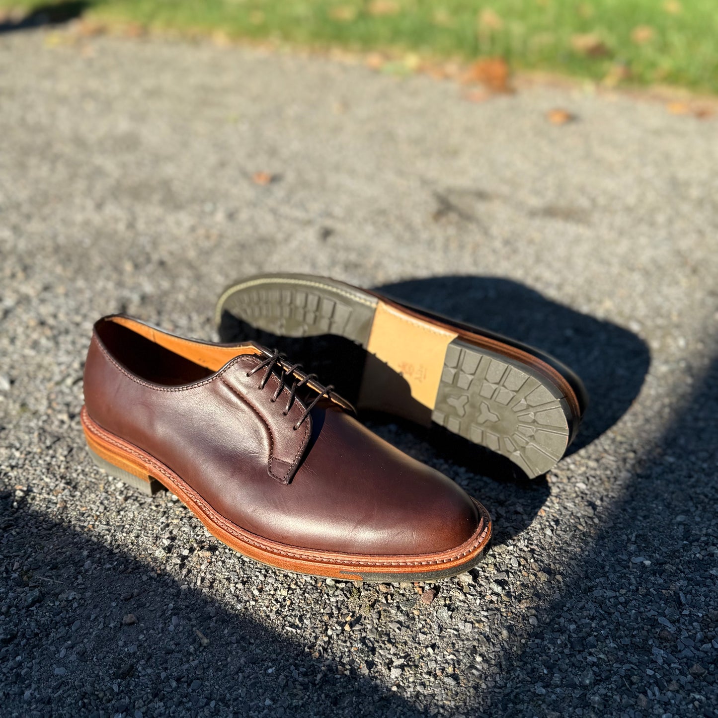 A pair of ALDEN SHOES 9506C - Plain Toe Blucher w Commando Brown CXL are displayed outdoors on gravel; one shoe stands upright and the other shows its sole, with grass in the background highlighting the Barrie Last design.