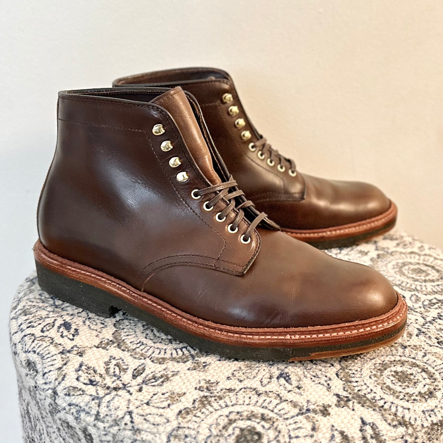 The Alden Madison D1848H Plain Toe Boot in brown Chromexcel leather, featuring metal eyelets and dark crepe rubber soles, rests on a patterned light fabric surface against a plain wall.