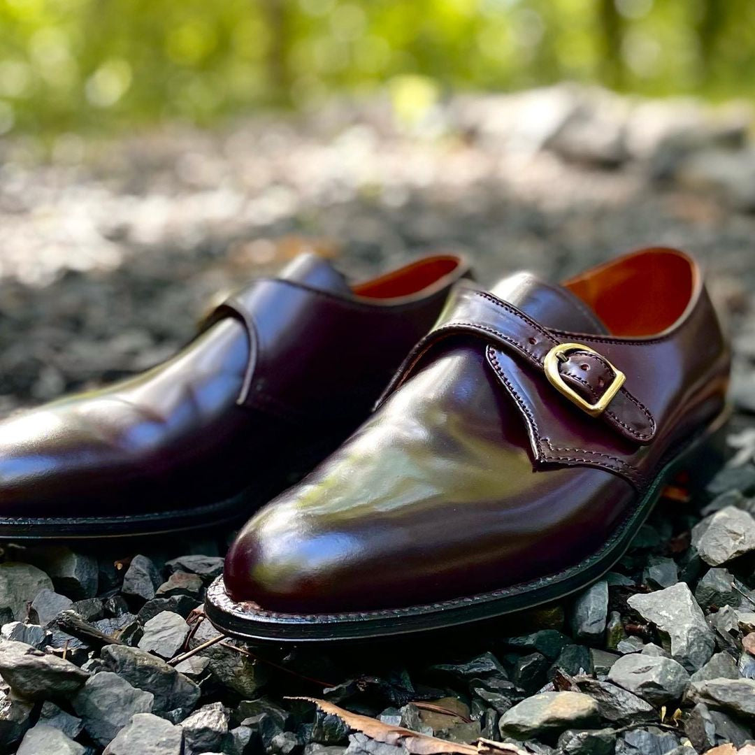 A pair of Alden Madison 954 - Monk Strap shoes in Color 8 Shell Cordovan with brass buckles rest on gray gravel. A blurred green backdrop suggests an outdoor setting, while the classic Aberdeen Last shape and leather sole ensure style and durability.
