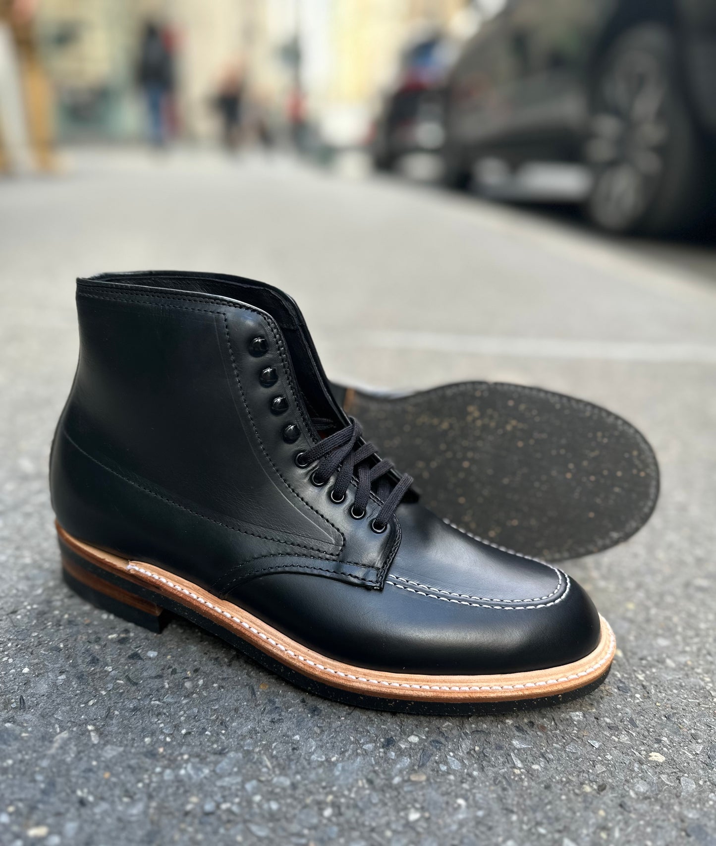 The Alden Madison 401 - Indy Boot in Black Chromexcel, featuring a tan welt and Neoprene cork sole, is shown on a city sidewalk—one boot upright and one on its side to highlight the durable sole construction.