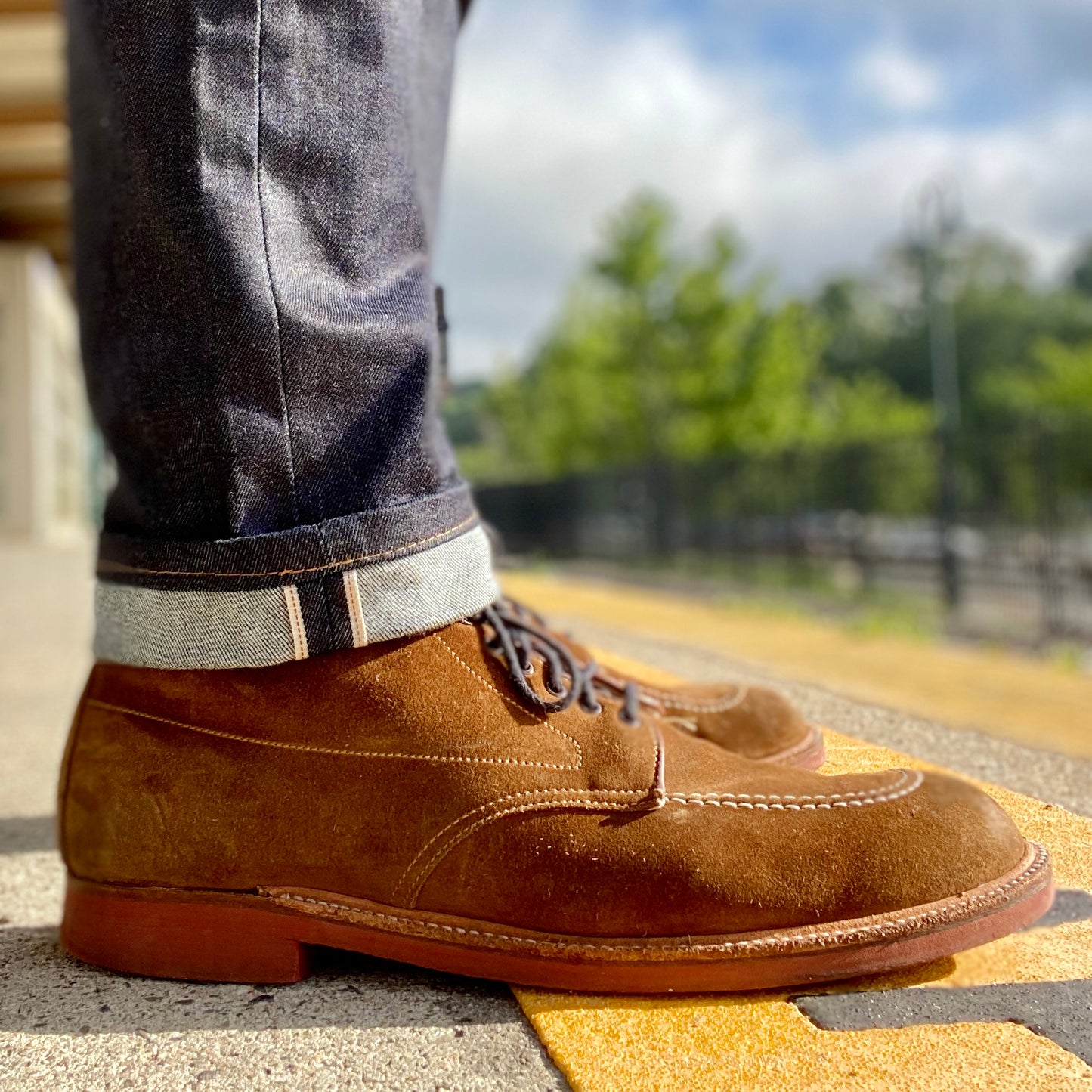 Close-up of a person wearing Alden Madisons 40531H Redwood Indy Boot in Snuff Suede, paired with dark denim jeans with rolled cuffs, standing on yellow tactile paving. The blurred background reveals greenery and a fence beneath a cloudy sky.