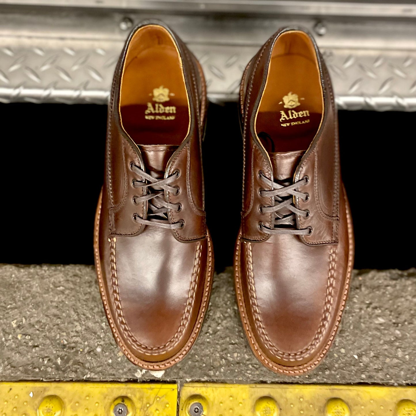 A top-down view displays a pair of D1611C - Ranger Mocc shoes from Alden Madison in Brown CXL leather with laces and Commando Sole, resting on a textured metal and rubber surface, showcasing the brand name and logo inside each shoe.