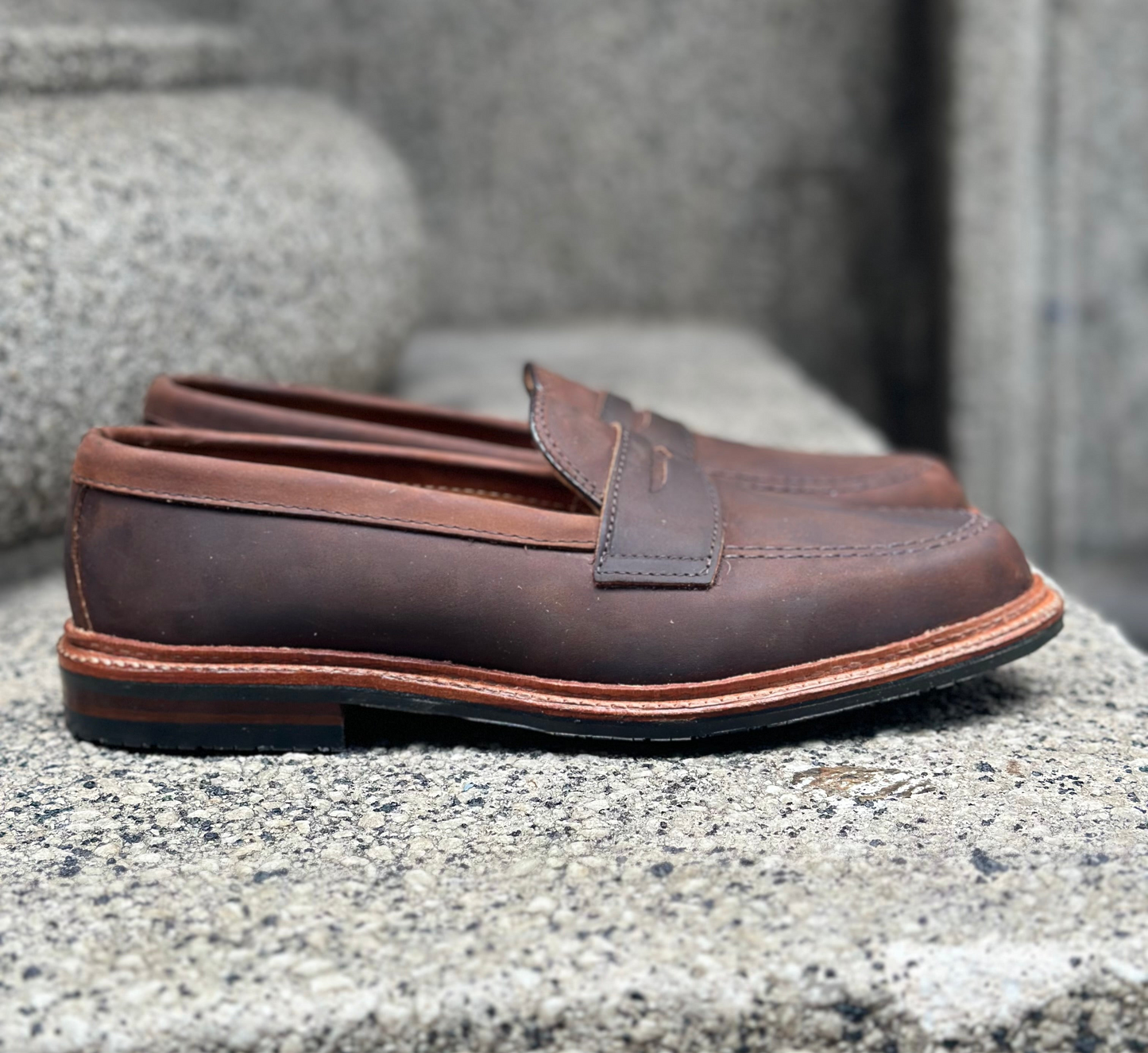 The D2216L - Smooth Tobacco Chamois Penny Loafer by Alden Madison showcases brown leather with an antique edge and polished finish, set on a textured stone background. Featuring a classic penny strap, the loafers have a tobacco smooth hue with a blurred backdrop emphasizing their sleek design.