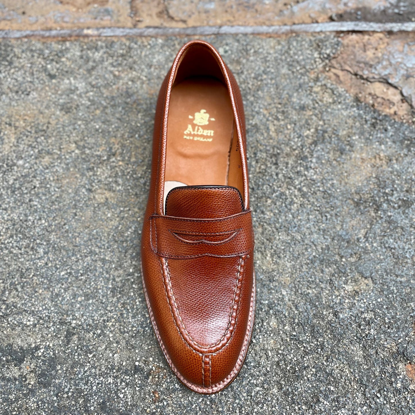 A single brown D1204 - Alpine Grain NST Loafer by Alden Madison, featuring visible stitching, penny slot, and Antique Flat Welt, rests on gray textured stone. The insole displays Alden New England in gold.