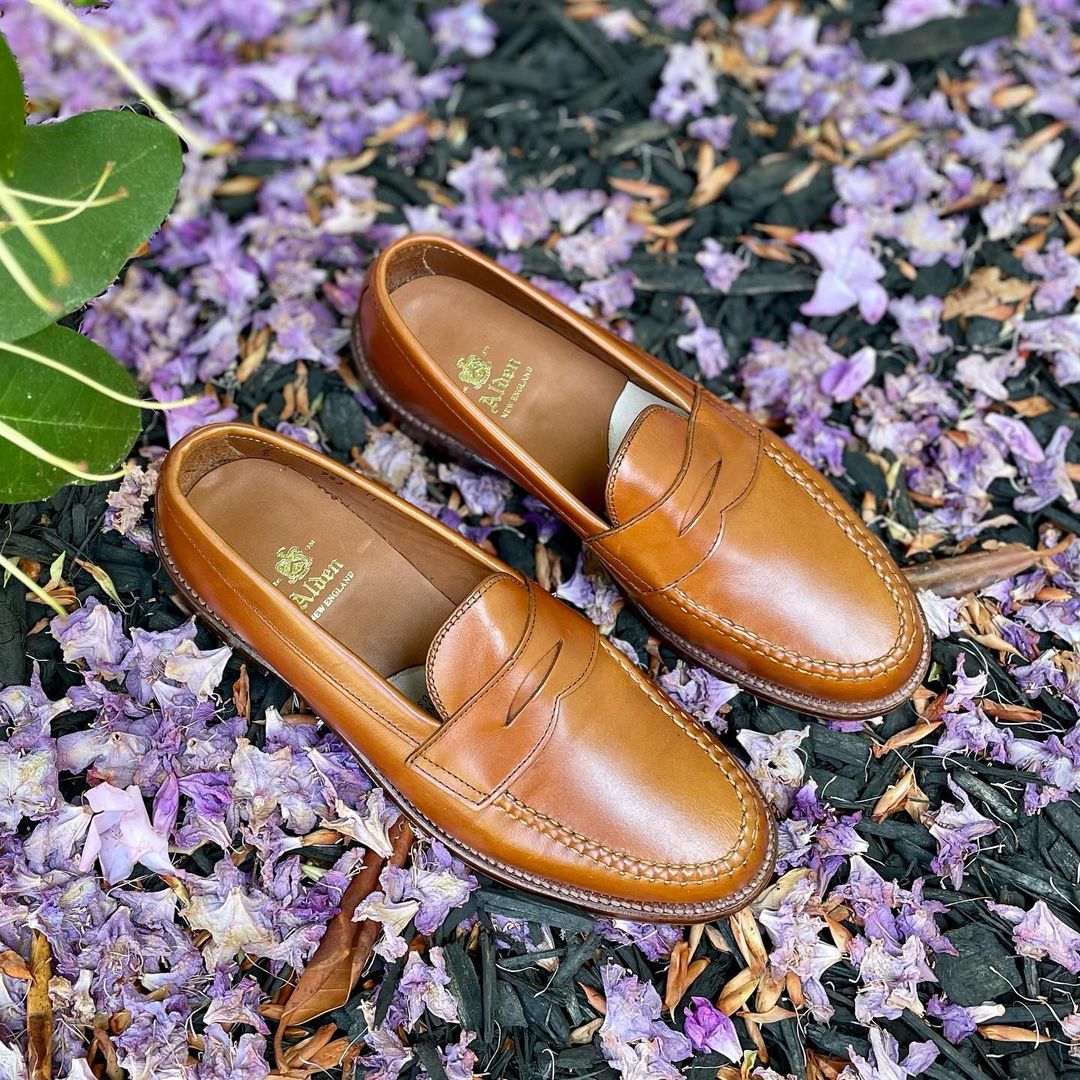 A pair of Alden Madisons 983 - Leisure Hand Sewn loafers in burnish tan calfskin rests on a purple flower and dark mulch. Featuring visible stitching and a branded logo on the insole, a large green leaf peeks from the top left corner to add a touch of nature.