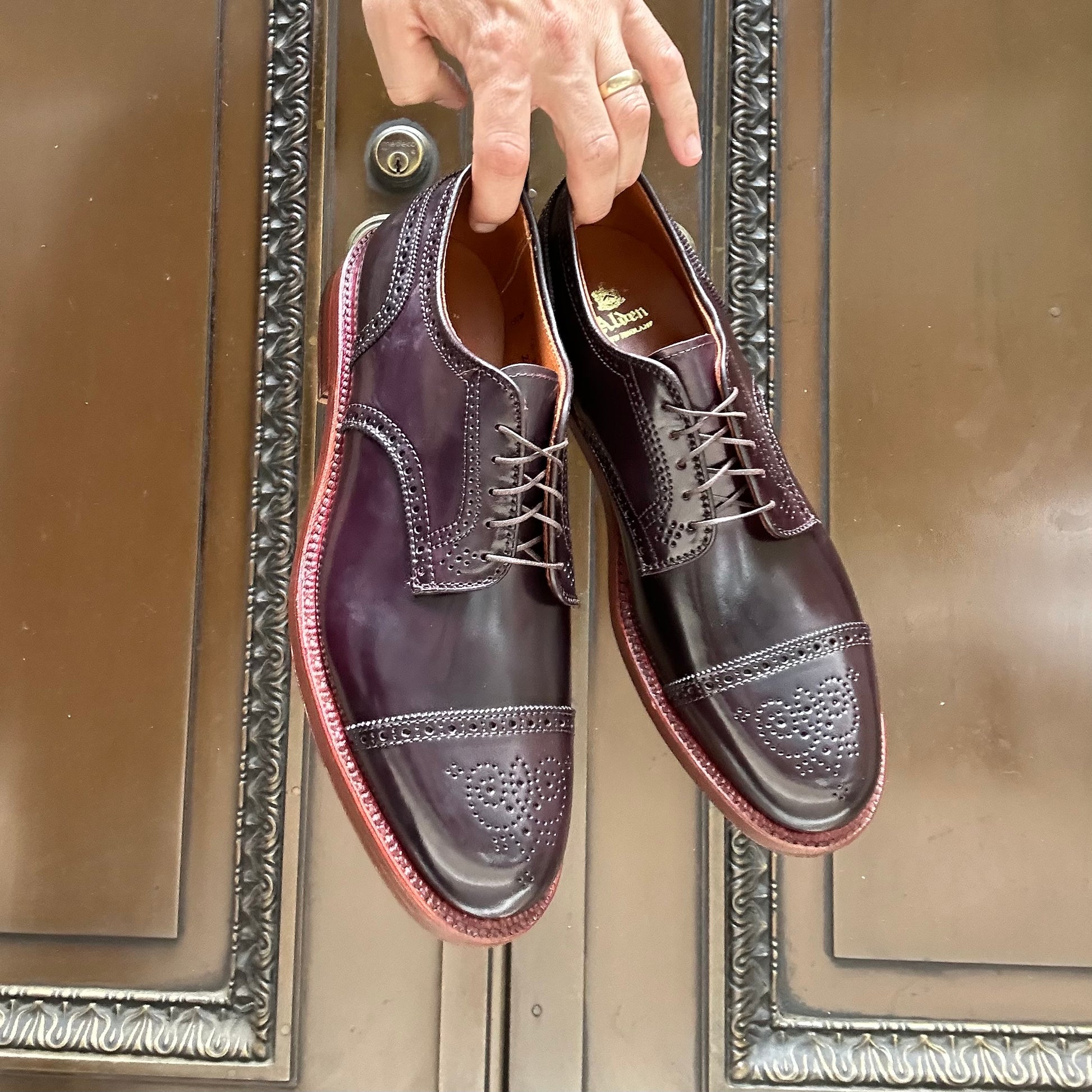 A hand holds the Alden Madison 21522 - Medallion Cap Toe Blucher Color 8, featuring elaborate brogue detailing in polished burgundy Shell Cordovan leather. The shoes are set against a decorative wooden door with ornate metal accents.