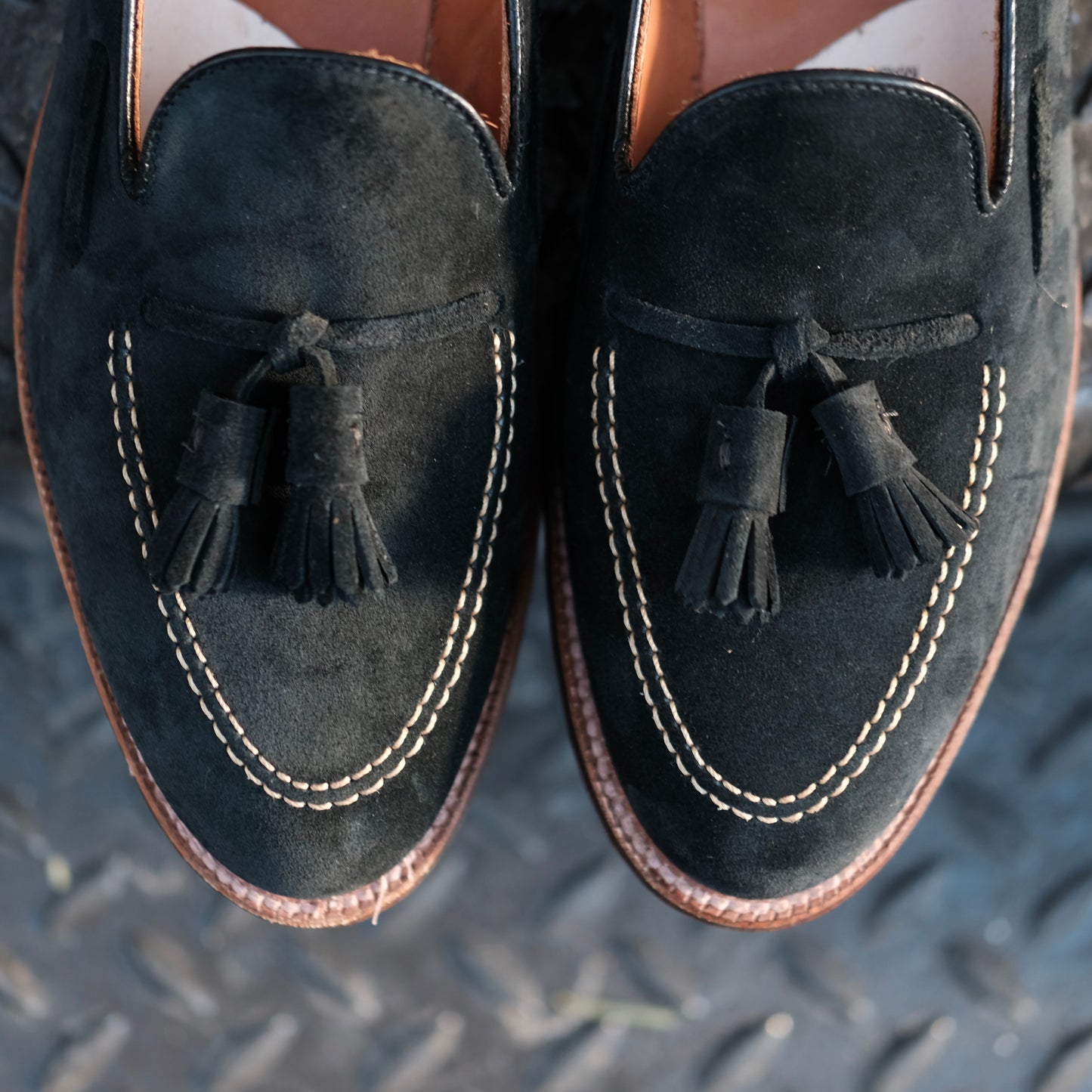 A close-up of the Alden Madison D0106 - Black Suede Tassel Loafer, showcasing white stitching around the toe, crafted on the Aberdeen Last and displayed on a textured dark surface.