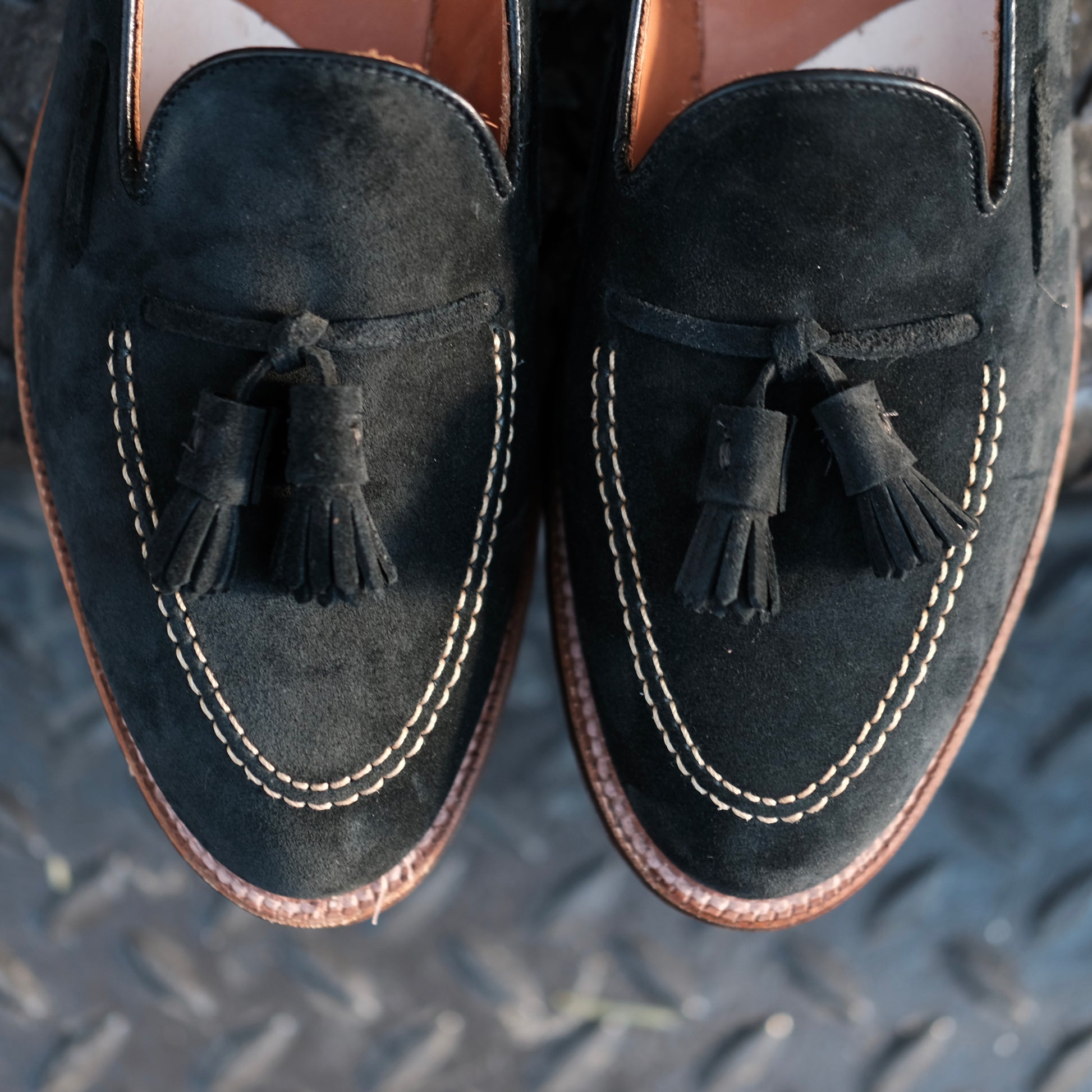 A close-up of the Alden Madison D0106 - Black Suede Tassel Loafer, showcasing white stitching around the toe, crafted on the Aberdeen Last and displayed on a textured dark surface.