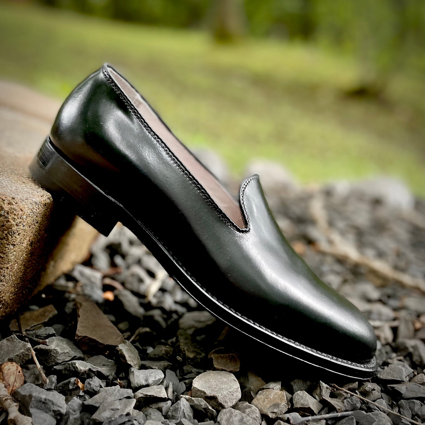 An 8815 Plain Toe Slip-On in Black Shell Cordovan by Alden Madison rests on its side among gray stones, set against a blurred green backdrop. The loafer, crafted on the Aberdeen Last, showcases a polished finish and low heel atop a raised edge.