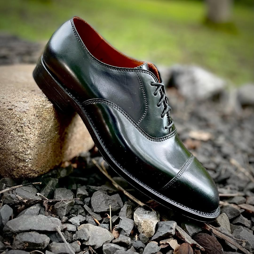 The Alden Madison 9071 - Straight Tip Bal in Black Shell Cordovan, a polished black lace-up dress shoe, is displayed on a stone amid dark gravel and rocks with blurred greenery in the background.