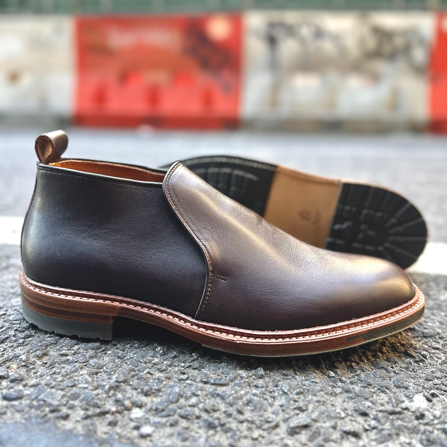 Alden Madison D2714C - 750 Slip On Chukka in Arabica Lux shown: one boot upright, the other on its side, displayed on textured outdoor pavement with a blurred red and white barrier in the background.