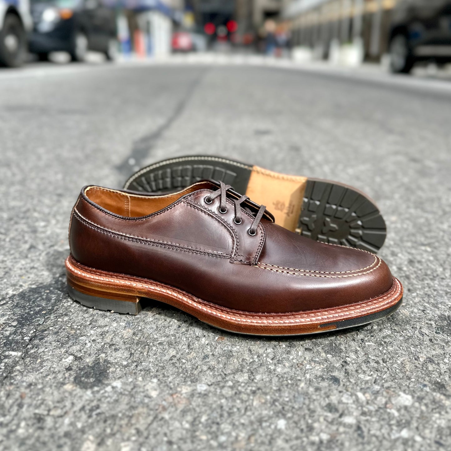 The Alden Madison D3616C - Ranger Mocc in Brown CXL lace-up shoes are showcased on a street. Highlighted by a Mocc Toe, Commando Sole, and detailed stitching, they feature a robust design set against a blurred urban background.