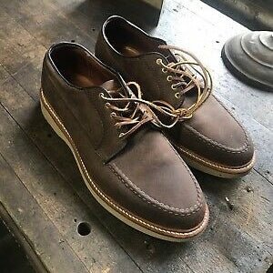 A pair of ALDEN SHOES 73980 - Tobacco Chamois Ranger Mocc with Wedge, featuring brown chamois leather, white stitching, and light wedge soles, displayed on a rustic wooden surface.