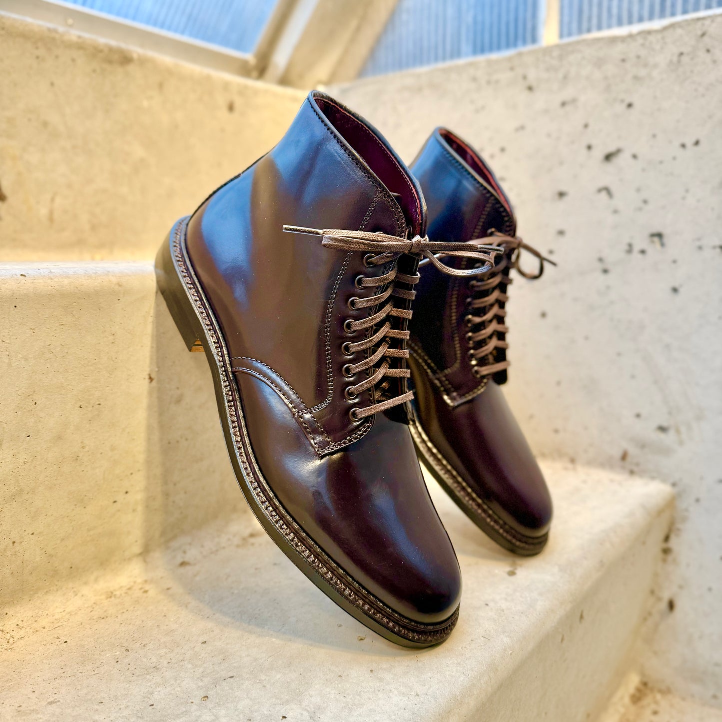 A pair of Alden Madisons 45168 - Plain Toe Director Boot in Color 8 Shell Cordovan made with shiny dark brown leather sits on white concrete steps, featuring a polished finish and thick soles crafted with a Single Oak Leather Sole, softly lit by daylight from the side.