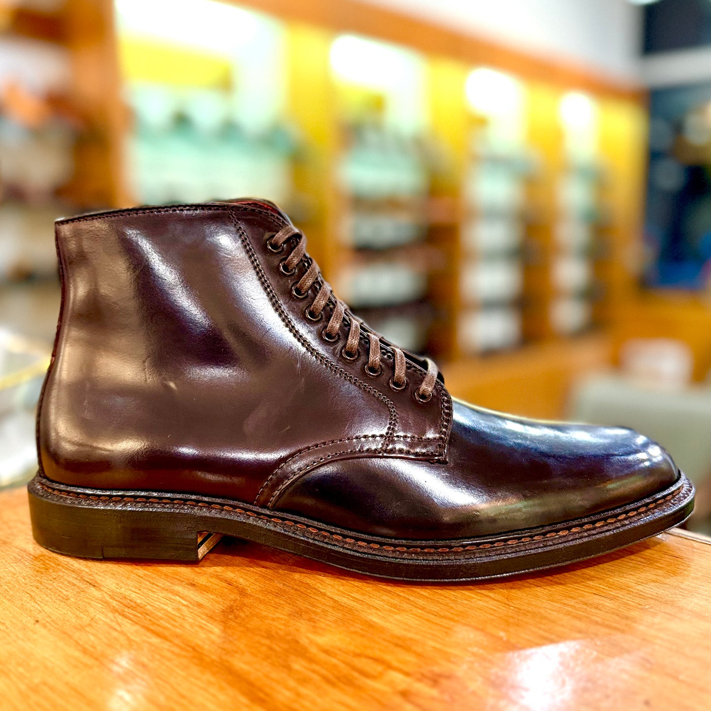 Displayed on a wooden surface, the Alden Madison product 45168 is a Plain Toe Director Boot in Color 8 Shell Cordovan, showcasing detailed stitching and laces. The blurred background highlights its glossy finish and refined Single Oak Leather Sole for superior durability.