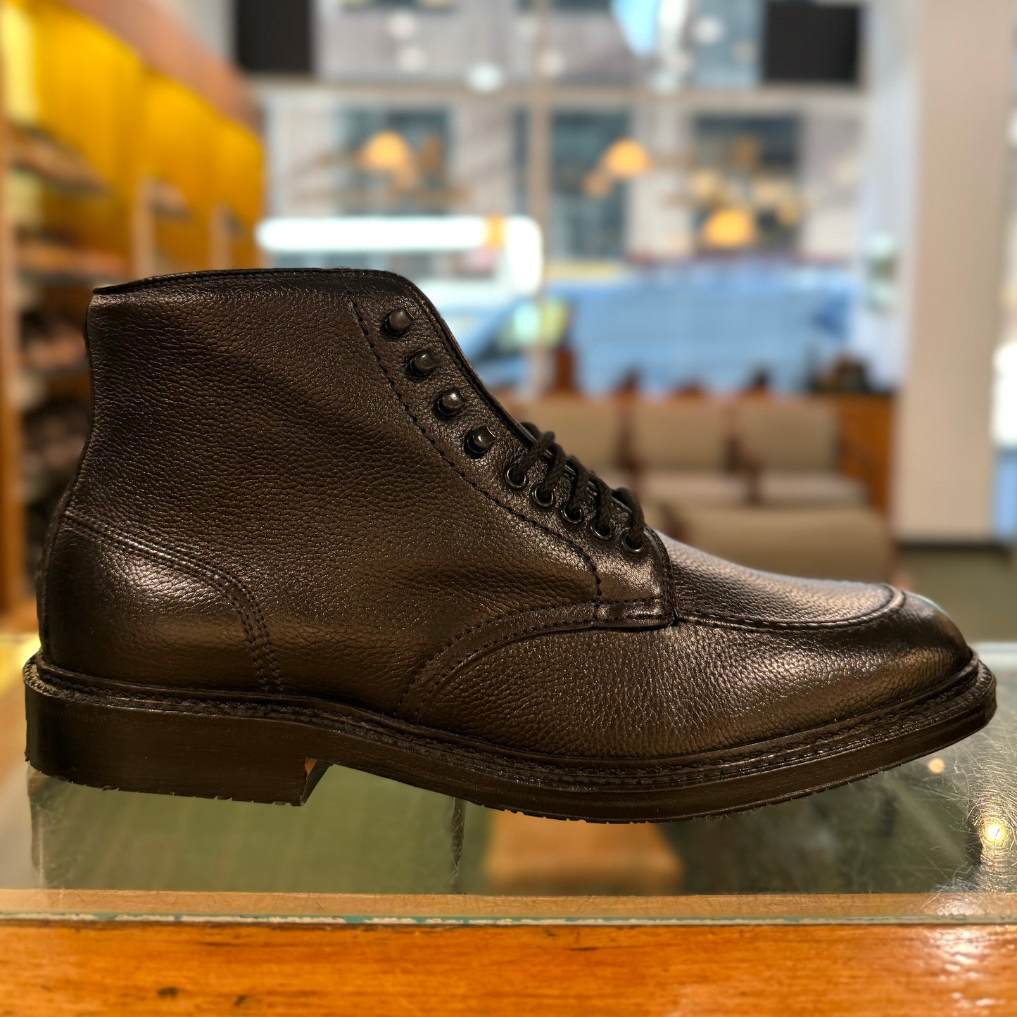 The Alden Madison 39615LH Han Boot in Black Regina Grain with a textured finish is shown on a glass surface in a well-lit store, with shelves and seating in the background.